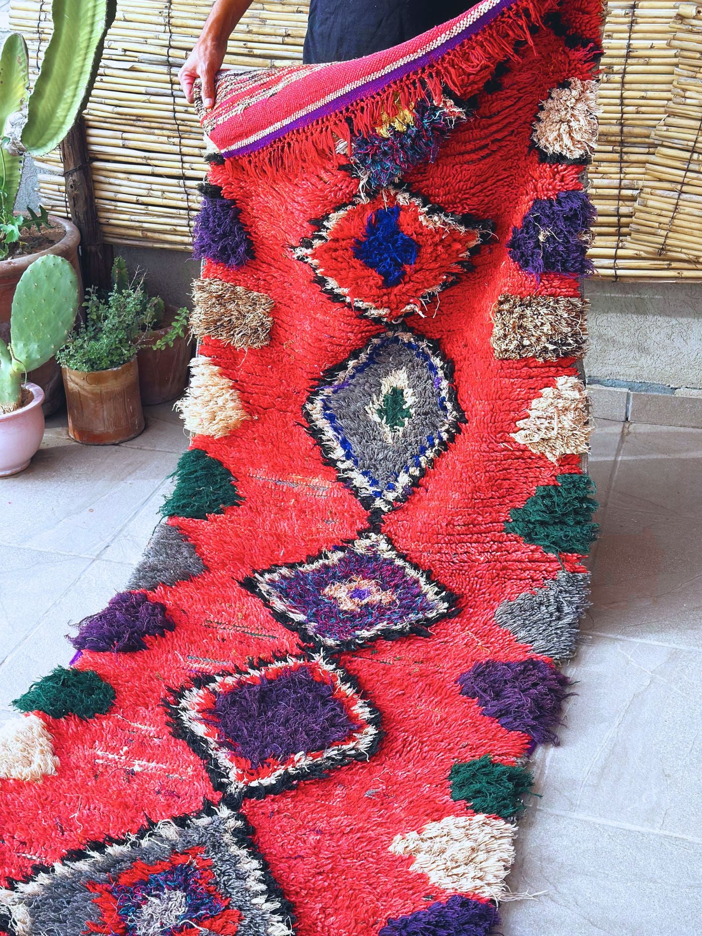 Handwoven Moroccan Boucherouite rug with vibrant geometric diamond motifs, featuring red, violet, green, and cream colors. Crafted by Berber artisans using sustainable recycled materials and wool, ideal for bohemian, eclectic, or modern interiors. Unique cultural design with symbolic motifs from the High Atlas Mountains of Morocco. Photo of full size rug width being held by a person, for size relativity. 