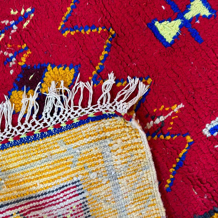 This handwoven Moroccan runner rug with bold red, yellow, and pink hues, featuring geometric Berber motifs is Perfect for boho, modern, contemporary or eclectic interiors. The motifs include diamond (symbolizing protection), zig zag mountain lines, and hourglass shaped butterflies. Photo shows tassels. 