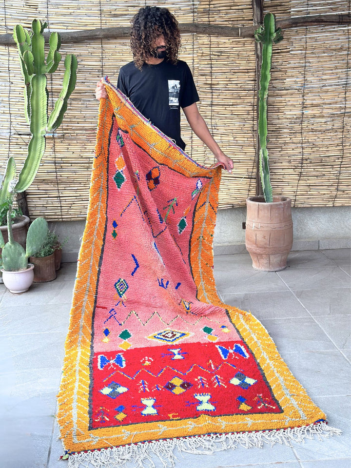 This handwoven Moroccan runner rug with bold red, yellow, and pink hues, featuring geometric Berber motifs is Perfect for boho, modern, contemporary or eclectic interiors. The motifs include diamond (symbolizing protection), zig zag mountain lines, and hourglass shaped butterflies. Photo shows someone holding rug for real life size and color. 